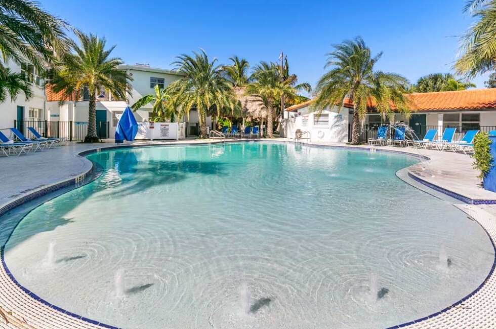 A large pool with fountains and lounge chairs surrounded by palm trees and buildings.