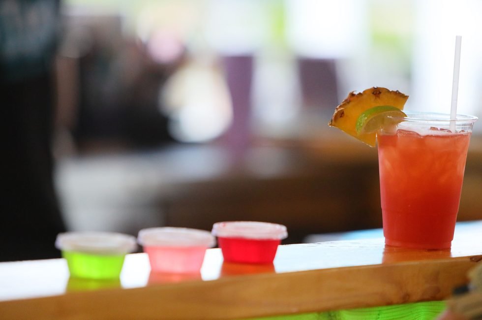 A red drink with a straw and fruit garnish next to three small cups of colorful drinks on a wooden table.