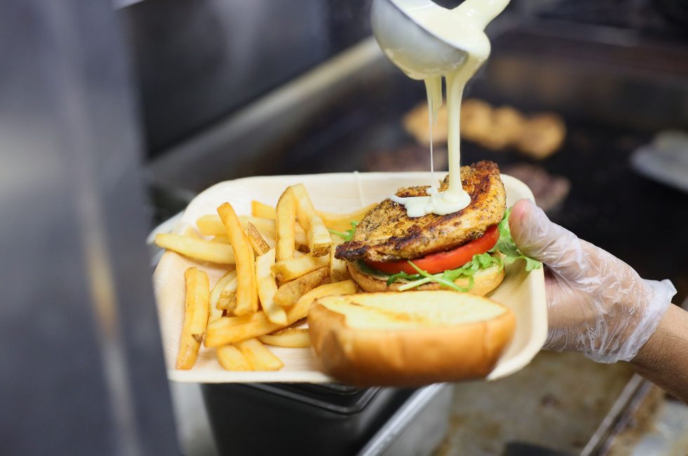 A hand in a clear glove holding a tray with a grilled chicken sandwich and fries, with sauce being poured over the sandwich.